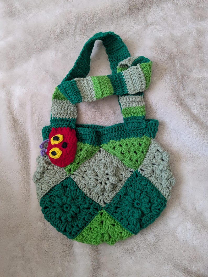 May include: A handmade crochet bag featuring a patchwork design in shades of green and gray. The bag has a green handle and a decorative caterpillar applique in red, yellow, and purple. The bag is approximately 10 inches wide.