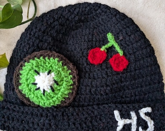 May include: Black crocheted beanie with a green kiwi and red cherries appliques. The beanie has the initials "HS" embroidered in white on the brim.