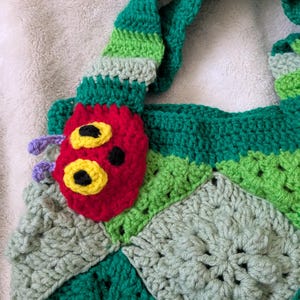 May include: A handmade crochet tote bag featuring a colorful caterpillar design. The bag is primarily green with a red caterpillar face, yellow eyes, and purple antennae. The bag's strap is striped green and light green.