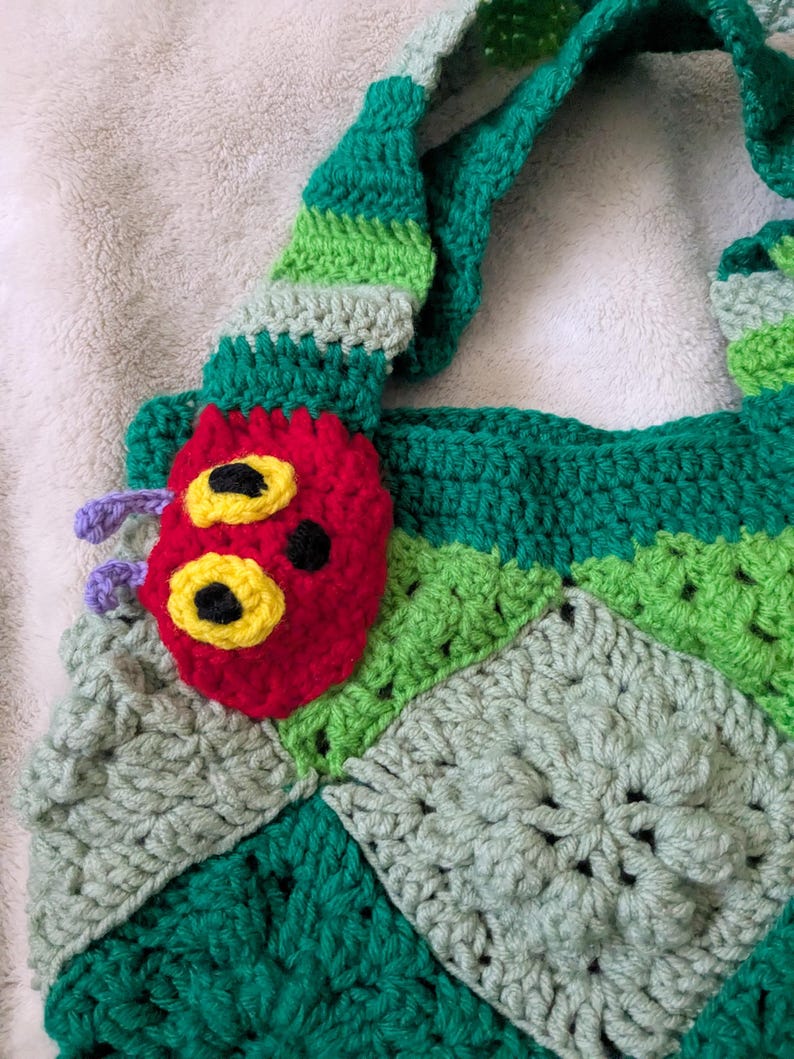 May include: A handmade crochet tote bag featuring a colorful caterpillar design. The bag is primarily green with a red caterpillar face, yellow eyes, and purple antennae. The bag's strap is striped green and light green.