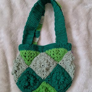 May include: A handmade crochet bag in shades of green and gray. The bag features a patchwork design with square motifs and a sturdy handle. The bag is made of yarn and is suitable for carrying items. The bag is a unique, handcrafted accessory.