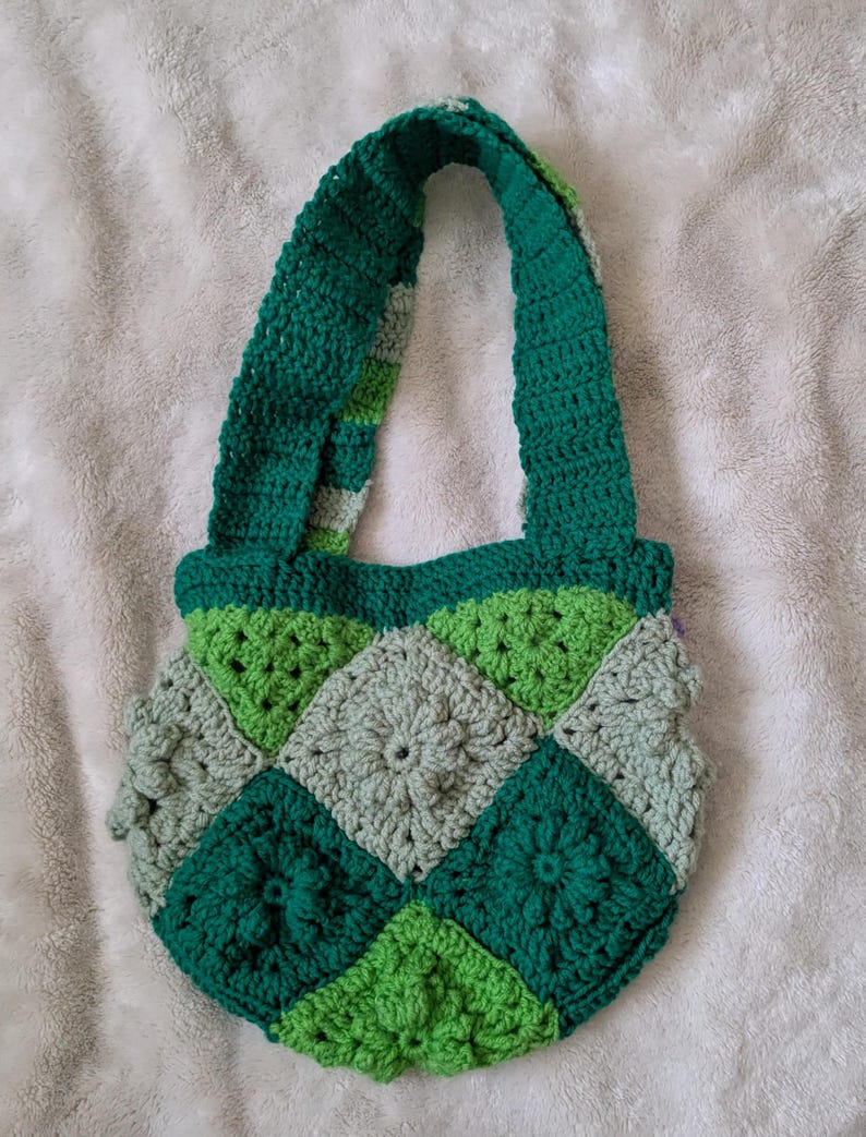 May include: A handmade crochet bag in shades of green and gray. The bag features a patchwork design with square motifs and a sturdy handle. The bag is made of yarn and is suitable for carrying items. The bag is a unique, handcrafted accessory.