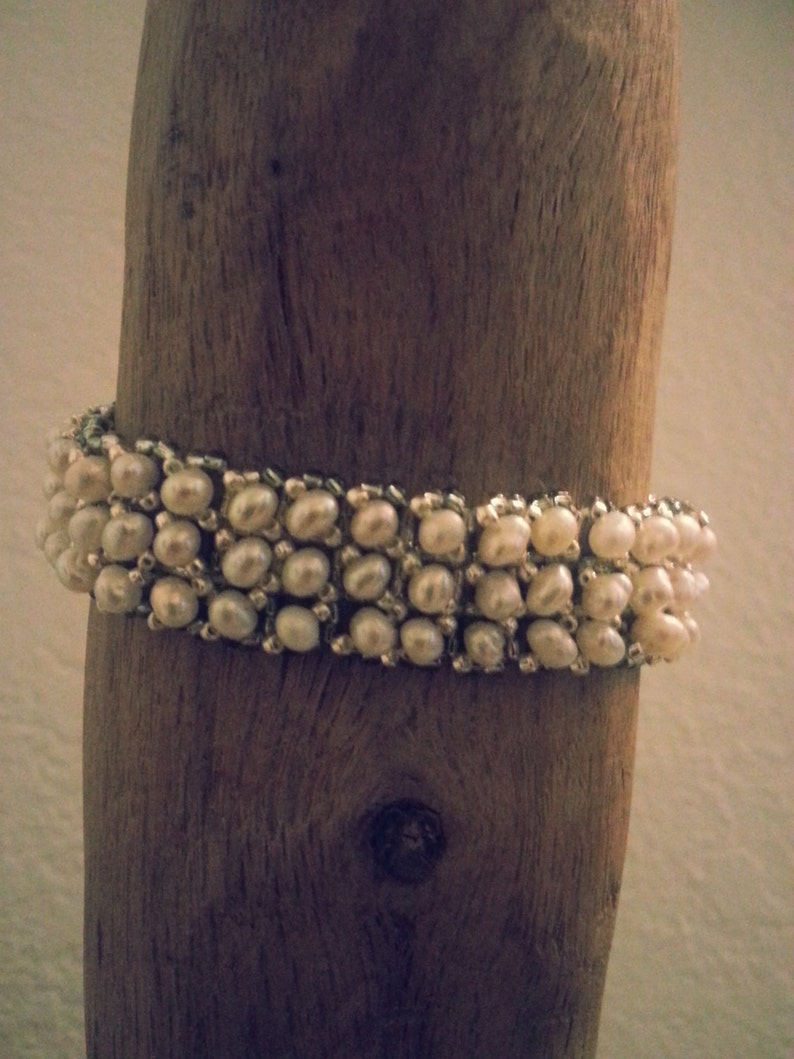 May include: A white pearl bracelet with a woven design. The bracelet is displayed on a piece of driftwood.