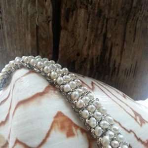May include: A white pearl bracelet with a silver chain. The bracelet is on a white seashell with brown markings.