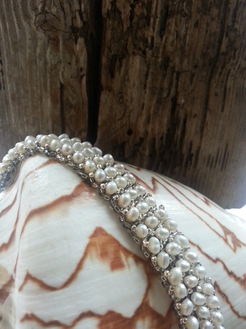 May include: A white pearl bracelet with a silver chain. The bracelet is on a white seashell with brown markings.