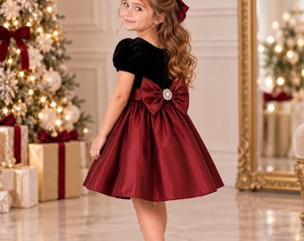 May include: A dress featuring a black velvet bodice with short puff sleeves and a deep red satin skirt. The waist is accented with a large red satin bow at the back, which is centered with a circular rhinestone embellishment. The skirt is knee-length.
