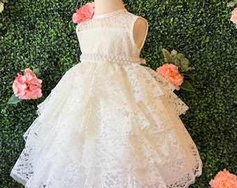 May include: A sleeveless ivory lace dress with a layered skirt, displayed on a mannequin. The dress features a pearl-beaded belt and a delicate lace overlay. The background includes pink flowers and a green hedge, creating a soft, elegant aesthetic.