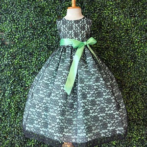 May include: A black lace dress with a light green underlay and a matching green ribbon sash. The dress is displayed on a mannequin against a green hedge backdrop. The dress has a round neckline and a full skirt.