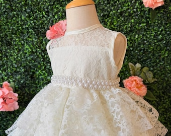 May include: Ivory lace dress with a pearl beaded belt. The dress features a layered skirt and a delicate lace bodice. The dress is displayed on a mannequin with a green hedge and pink flowers in the background.