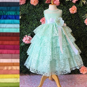 May include: A mint green tiered lace dress with a white satin ribbon and flower accent. The dress is displayed on a wooden mannequin, set against a backdrop of pink flowers and green foliage. A color swatch of fabric samples is on the left.