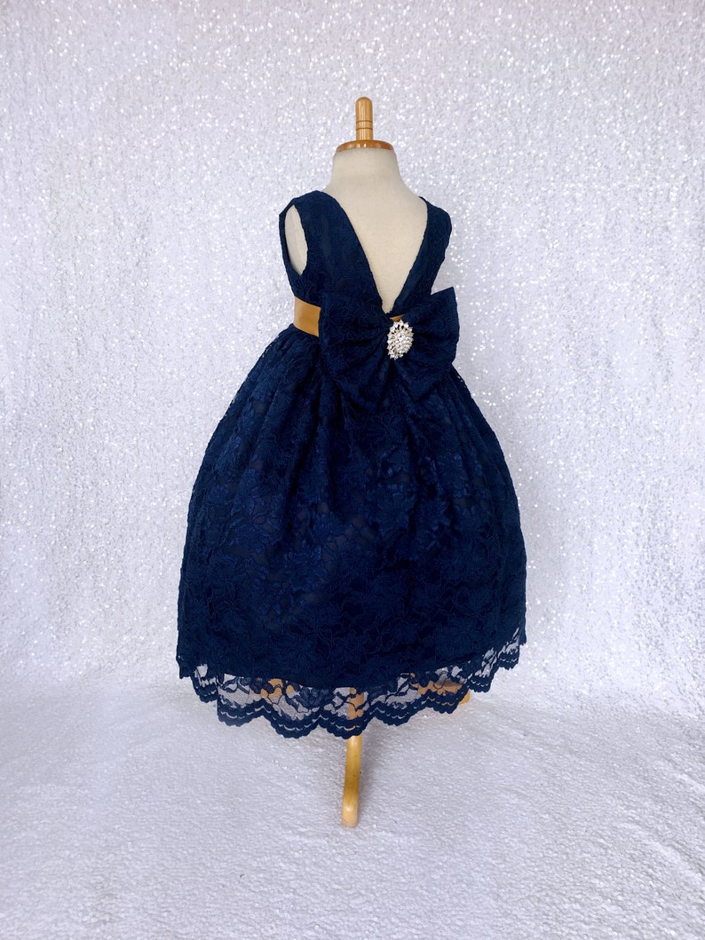 May include: A navy blue lace dress with a gold sash and a large bow in the back. The dress has a scalloped hem and a v-shaped neckline.
