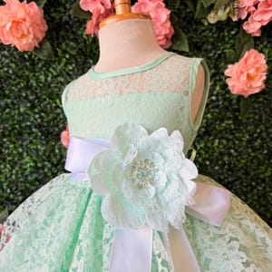 May include: A mint green lace dress for a child, displayed on a mannequin. The dress features a round neckline, a white satin sash, and a large flower embellishment with a rhinestone center. The background is a wall of pink flowers and green foliage.