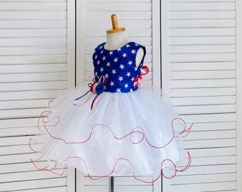 Op de afbeelding: Een witte tule jurk met rode bies en een blauw en wit sterrenpatroon op het lijfje. De jurk is versierd met rode, witte en blauwe linten.