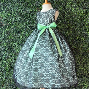 May include: A sleeveless dress on a mannequin, featuring a black lace overlay over a light blue fabric. The dress has a full skirt and a light green satin ribbon tied in a bow at the waist. The dress is trimmed with black lace.