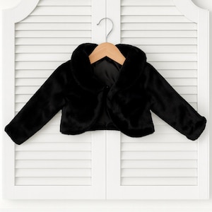 May include: A black faux fur bolero jacket with a rounded collar and long sleeves. The jacket is hanging on a wooden hanger against a white shuttered door. The bolero is a cropped style, designed to be worn over other garments.