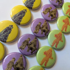 May include: A collection of round buttons with different designs. The buttons are yellow, purple, and green. The designs include a crown, a pair of figures, and a single figure.