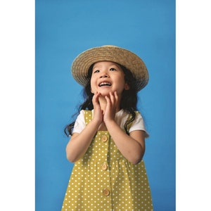 May include: A young girl wearing a yellow polka dot dress with white buttons and a straw hat, looking up with a smile against a blue background.