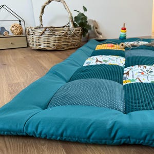 A cozy quilt with a blue and white patterned patchwork, placed on a wooden floor. In the background, there is a wicker basket, a toy, and a plant.