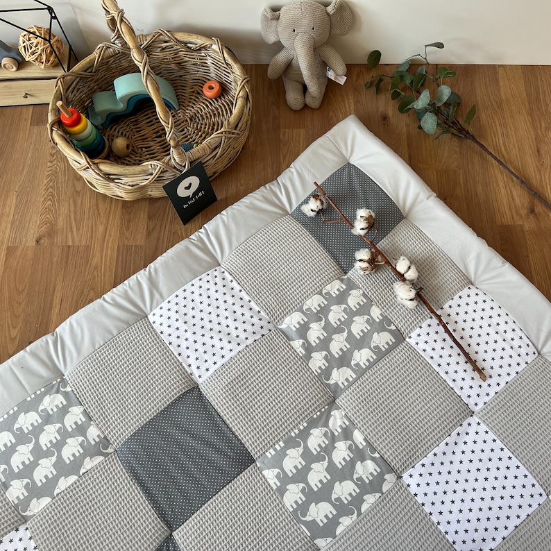 a cozy and inviting baby's room, with a gray and white quilt-like bedspread, a wicker basket filled with toys, and a stuffed elephant toy. The room has a warm and homely atmosphere, with a wooden floor and a white wall as the backdrop.