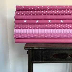 May include: A stack of eight pink fabric rolls with different white patterns, including polka dots, stars, and stripes. The fabric rolls are stacked on a dark wooden table.