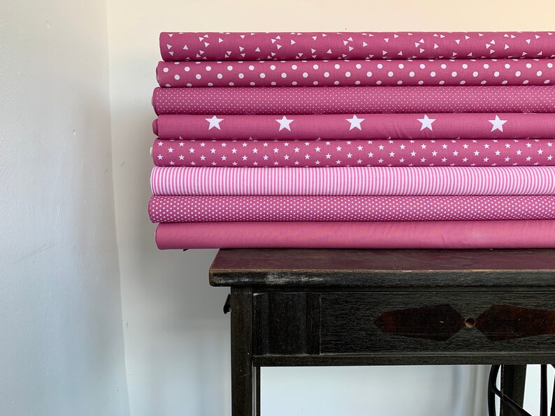 May include: A stack of eight pink fabric rolls with different white patterns, including polka dots, stars, and stripes. The fabric rolls are stacked on a dark wooden table.