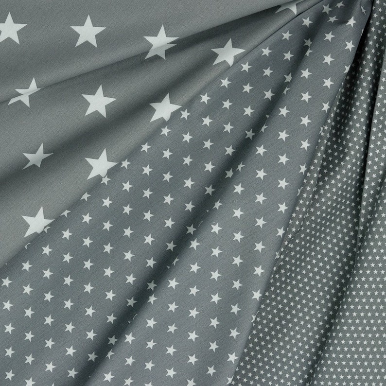 May include: Gray fabric with a white star pattern. The fabric is folded, showing different densities of star arrangements, from large, spaced-out stars to a dense field of small stars. The fabric has a soft, flowing appearance.