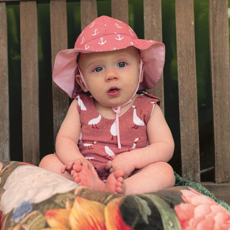 K&ouml;nnte beinhalten: Ein Baby tr&auml;gt einen rosa Sonnenhut mit wei&szlig;en Anker-Mustern und einen passenden rosa Strampler mit wei&szlig;en G&auml;nse-Illustrationen. Das Baby sitzt auf einem floral gemusterten Kissen auf einer Holzbank.