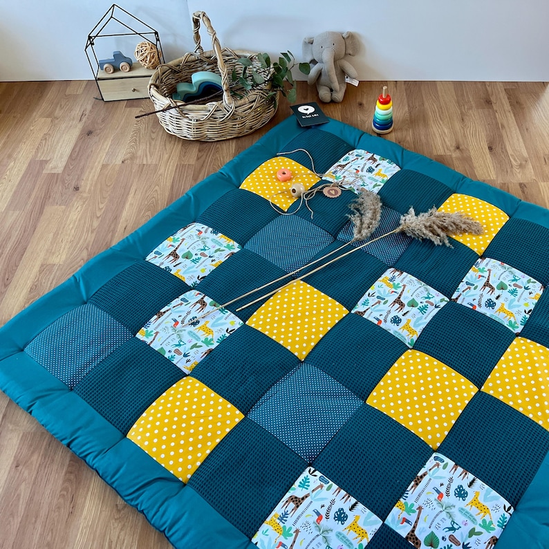 a colorful quilt with a patchwork of blue, yellow, and white squares, arranged on a wooden floor. There are also various toys and decorative items scattered around the quilt, including a basket, a stuffed elephant, and a toy car.