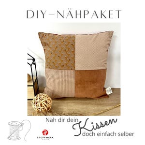 May include: DIY sewing kit for a cushion with a patchwork design. The cushion is made of four different fabrics: a brown fabric with a pattern of arches, a light brown fabric with a subtle texture, a light brown waffle fabric, and a brown corduroy fabric. The cushion is sitting on a wooden surface with a ball of twine and a tag that says "Näh dir dein Kissen doch einfach selber".
