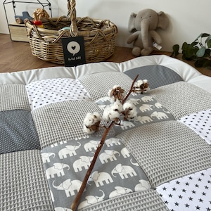 a cozy and charming nursery scene, with a gray and white elephant-themed quilt, a wicker basket, and a stuffed elephant toy. The quilt has a patchwork design with gray and white elephants, and a branch with white cotton balls rests on top of it. The basket is filled with various items, and the stuffed elephant toy is placed next to it.