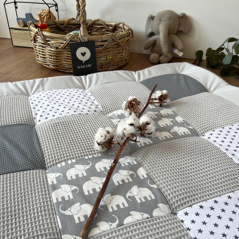 a cozy and charming nursery scene, with a gray and white elephant-themed quilt, a wicker basket, and a stuffed elephant toy. The quilt has a patchwork design with gray and white elephants, and a branch with white cotton balls rests on top of it. The basket is filled with various items, and the stuffed elephant toy is placed next to it.
