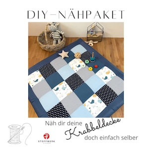 Könnte beinhalten: Ein Patchwork-Spielteppich für Babys mit blauen, grauen und weißen Quadraten. Einige Quadrate weisen Wal- und Tupfenmuster auf. Der Teppich ist von einem dunkelblauen Rand umgeben. Der Text oben lautet "DIY-NÄHPÄKET" und unten "Näh dir deine Krabbeldecke doch einfach selber."