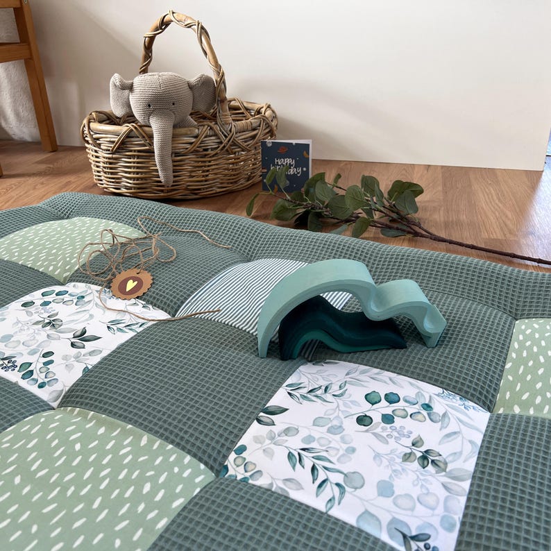 a cozy and inviting scene with a quilted green and white bedspread, a wicker basket, a stuffed elephant toy, and a greeting card on the floor.