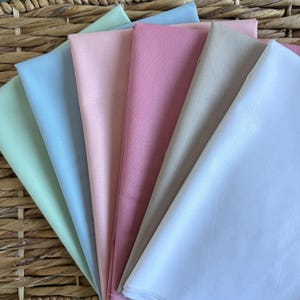 Puede incluir: Una colección de servilletas de tela dobladas en varios colores pastel, incluyendo verde claro, azul, rosa y blanco. Las servilletas están dispuestas en forma de abanico, mostrando su textura suave y variaciones de color. El fondo es una cesta tejida.