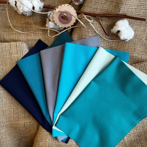 Könnte beinhalten: Eine Sammlung von acht Stoffproben in verschiedenen Blautönen und Grüntönen, die fächerförmig auf einem Jute-Hintergrund angeordnet sind. Die Proben sind glatt und haben einen dezenten Glanz.