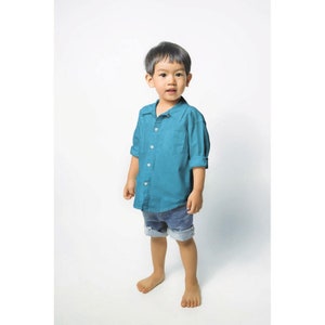 May include: A young boy wearing a teal blue button-down shirt and denim shorts stands in front of a white background.