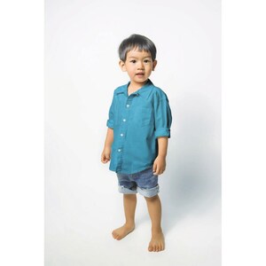 May include: A young boy wearing a teal blue button-down shirt and denim shorts stands in front of a white background.