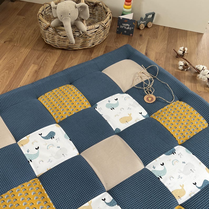 a cozy and colorful quilt with a patchwork design, placed on a wooden floor. The quilt has a mix of blue, yellow, and white colors, with various animal patterns such as elephants, dolphins, and rabbits. There is also a basket with a stuffed elephant toy and some toys, including a toy car and a toy train, scattered around the room.