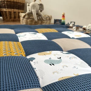 a close-up view of a quilt with a patchwork design, including a blue and white geometric pattern, a yellow and white pattern, and a blue and white pattern with a whale design. The quilt is placed on a bed, and there is a basket and a stuffed elephant toy visible in the background.