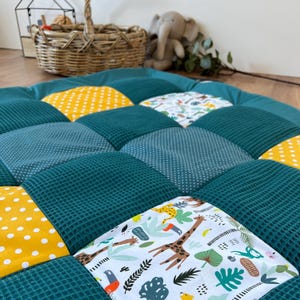a colorful quilt or mat with various patterns and textures, including polka dots, stripes, and animal prints, arranged in a patchwork design. There is a basket and a stuffed elephant toy visible in the background, adding to the cozy and playful atmosphere of the scene.