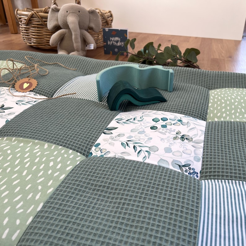 a cozy and colorful quilt with a variety of patterns and textures, including a green and white checkered pattern, a floral print, and a striped pattern. There is also a stuffed elephant toy and a greeting card with the message "Happy Birthday" on the floor.