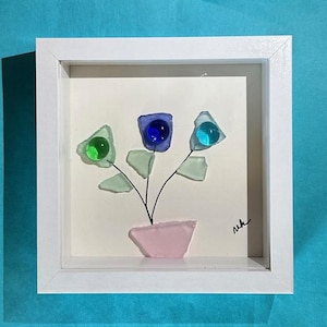 May include: A framed artwork featuring a floral arrangement crafted from sea glass. The flowers have green, blue, and purple glass petals with a pink glass vase. The artwork is set against a cream background within a white frame.