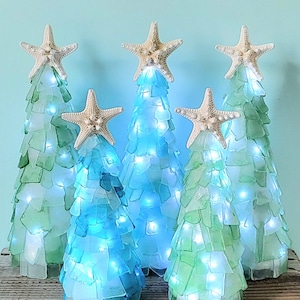 Puede incluir: Cinco árboles de Navidad decorativos hechos de vidrio marino en tonos azules y verdes. Cada árbol está coronado con una estrella de mar e iluminado con luces blancas. Los árboles están dispuestos sobre una superficie de madera contra un fondo azul claro.