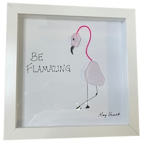 May include: White framed artwork with a pink flamingo made of glass and the text "BE FLAMAZING" in black. The artwork is signed "Mayji Kreut" in the bottom right corner.