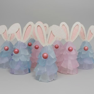May include: A group of Easter bunny decorations. The bunnies have white glitter ears with pink interiors and a pink sphere for a nose. The bodies are made of layered, translucent blue and pink material.