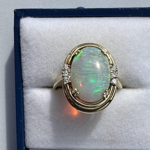May include: Oval opal ring with a gold band and accents. The opal has a play of color with green and blue hues. Small diamonds are set on either side of the opal.