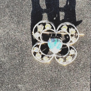 May include: A silver brooch with a blue-green gemstone in the center and a clover-shaped design with clear rhinestones.