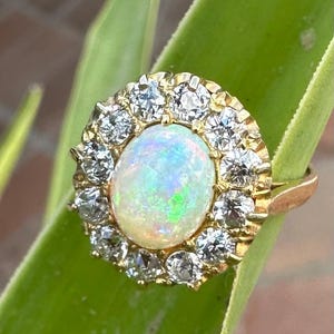 May include: A gold ring featuring a large, oval opal gemstone surrounded by a halo of small, clear diamonds. The opal displays iridescent colors. The ring is set on a green leaf.