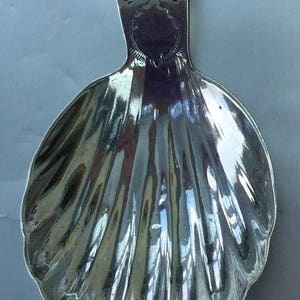 Puede incluir: Un plato de plata en forma de concha con un mango decorativo. El mango tiene un diseño intrincado de espirales y líneas.