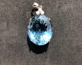 May include: A large, oval-shaped, light blue gemstone pendant with a silver setting. The pendant has three smaller, round, clear gemstones set in the top of the bail.
